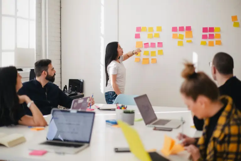 A woman is leading a meeting, pointing at colorful sticky notes on a whiteboard, while four colleagues seated at a table with laptops and notebooks listen attentively in a bright office setting. and speack also about event app for universities