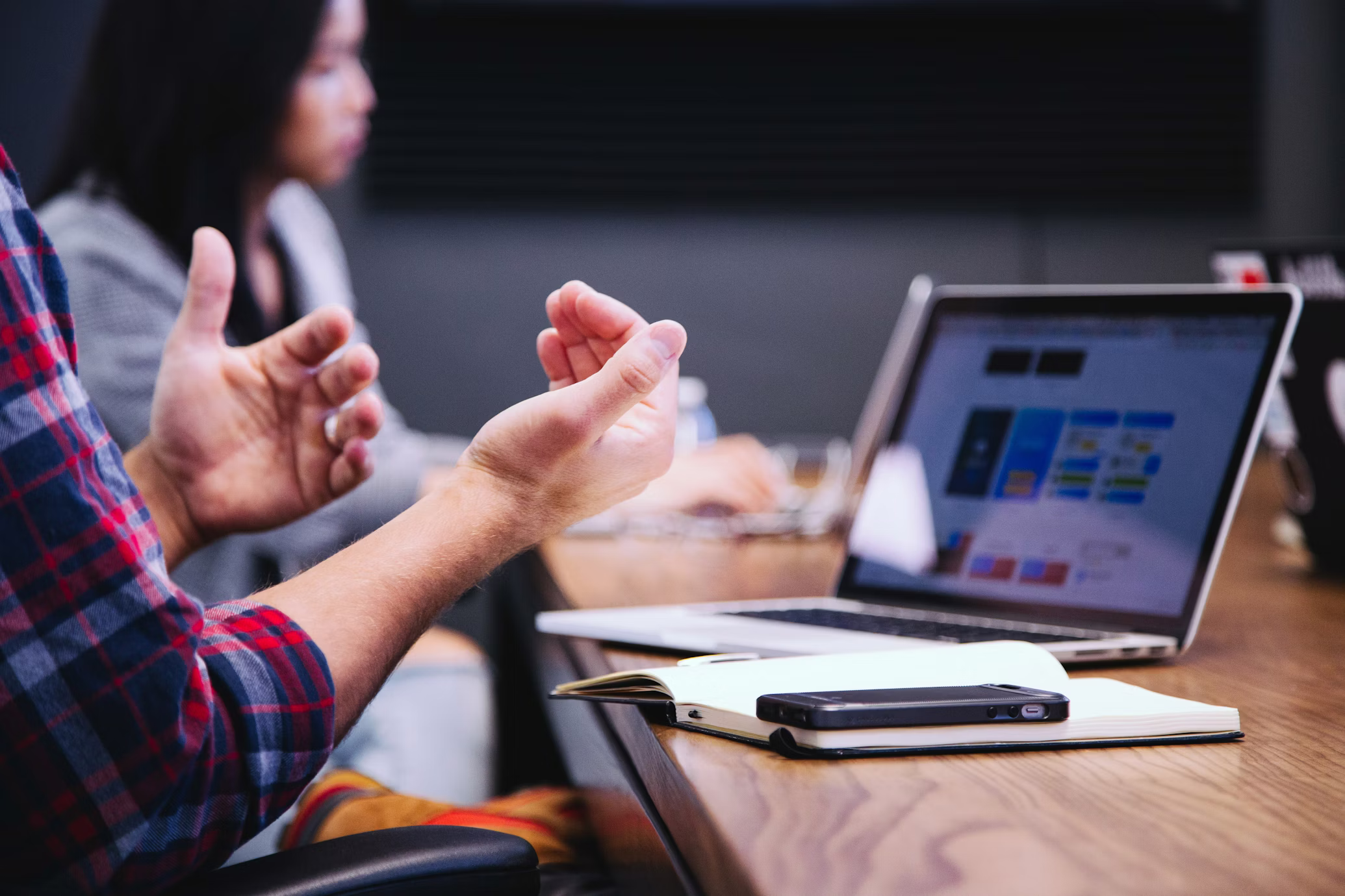 A person gestures with their hands while sitting at a wooden table, with a laptop displaying a colorful screen open in front of them. A smartphone and notebook lie nearby, and another person is visible in the background, slightly out of focus. for corporate event app