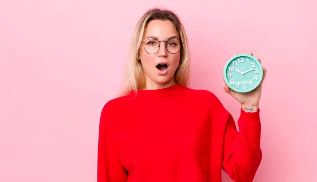 A surprised woman in a red sweater holds a turquoise clock with white numbers against a pink background. last-minute schedule changes